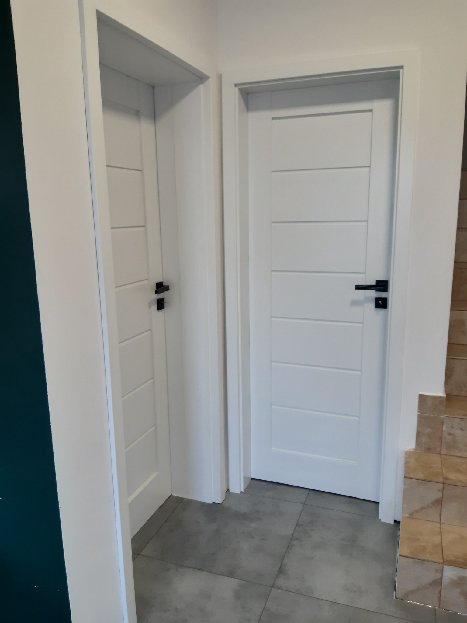 Dwa białe, nowoczesne drzwi wewnętrzne z poziomymi panelami i czarnymi klamkami, widoczne na tle szarej podłogi i fragmentu ściany z płytkami.