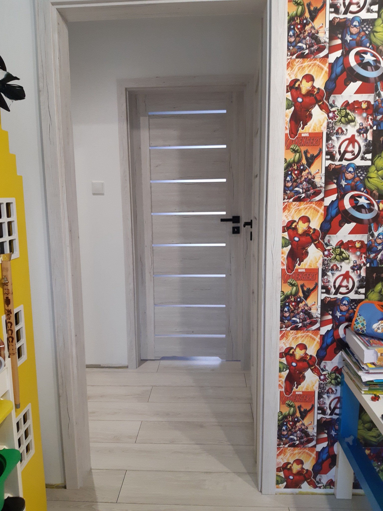 Wnętrze pokoju dziecięcego z drewnianą podłogą, białą ścianą z włącznikiem światła oraz drzwiami wewnętrznymi z poziomymi przeszkleniami i czarną klamką, obok ściana pokryta tapetą z motywem Avengers.