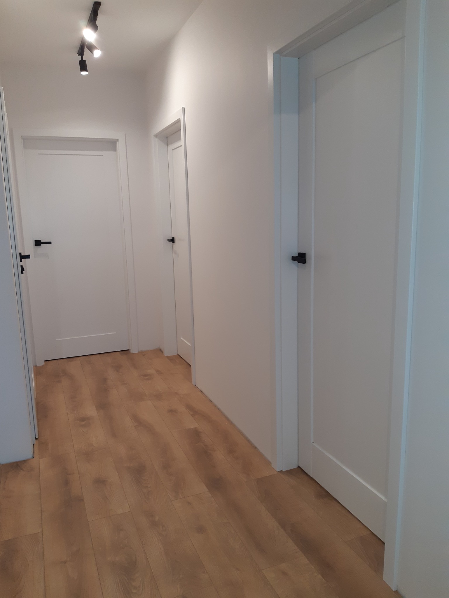 Korytarz z trzema białymi drzwiami z czarnymi klamkami i laminowaną podłogą w odcieniach brązu, oświetlony reflektorami sufitowymi.