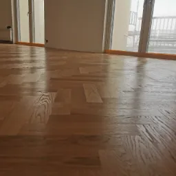 Świeżo ułożony parkiet w jodełkę, widoczne refleksy światła na lakierowanej powierzchni, okna z widokiem na rusztowania budowlane.