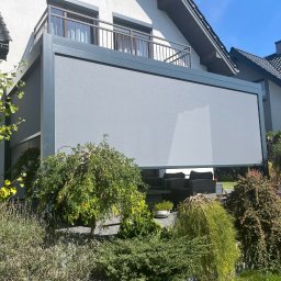 GRUPA ELWA KRZYSZTOF WAJDA - Szara, nowoczesna zabudowa balkonu z roletami przeciwsłonecznymi, widok z ogrodu. W tle biały dom z balkonem i ciemnym dachem. Uwagę przykuwają dekoracyjne krzewy.