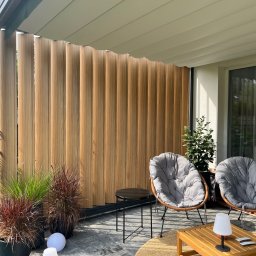 GRUPA ELWA KRZYSZTOF WAJDA - Nowoczesna pergola z drewnianymi lamelami, tworząca zacienioną przestrzeń na balkonie. Dwa fotele z poduszkami i stoliki kawowe uzupełniają aranżację.