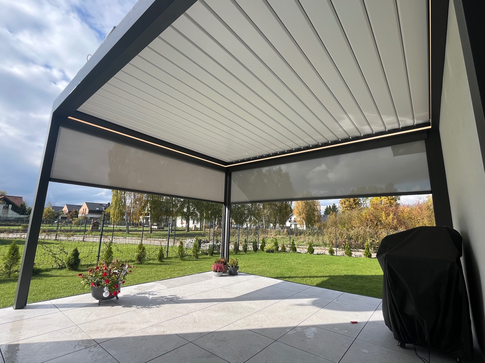 Nowoczesna, metalowa pergola z roletami przeciwsłonecznymi i oświetleniem LED, widok z dołu na taras z płytkami i zielony ogród w tle.