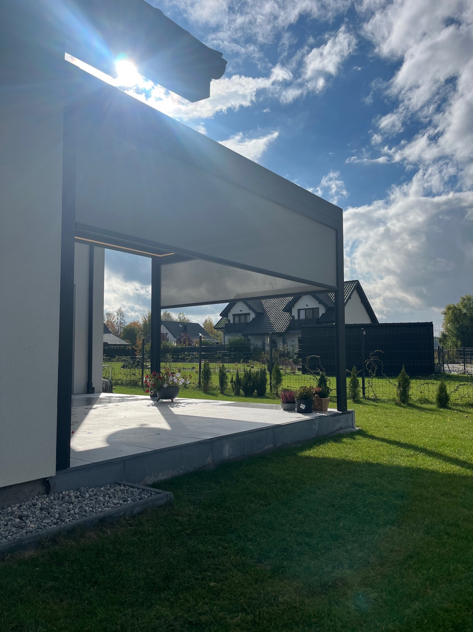 Nowoczesna konstrukcja zadaszenia tarasu z widokiem na ogród w Tarnowie. Minimalistyczny design, szara kolorystyka, rośliny w donicach. Słoneczny dzień.