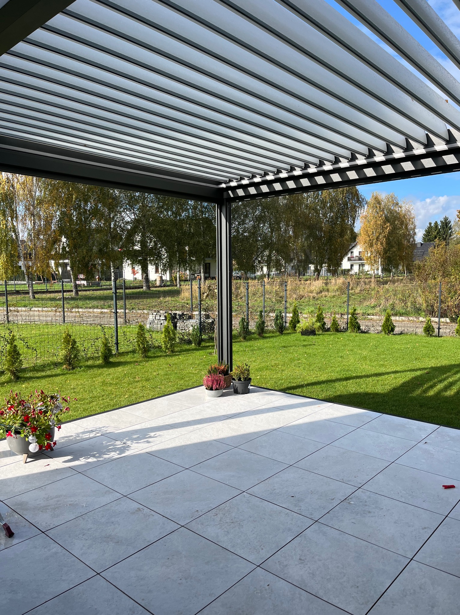 Nowoczesna, metalowa pergola z regulowanymi lamelami, zacieniająca taras z jasnych płytek. Widok na zadbany ogród z niskimi tujami i drzewami w tle.