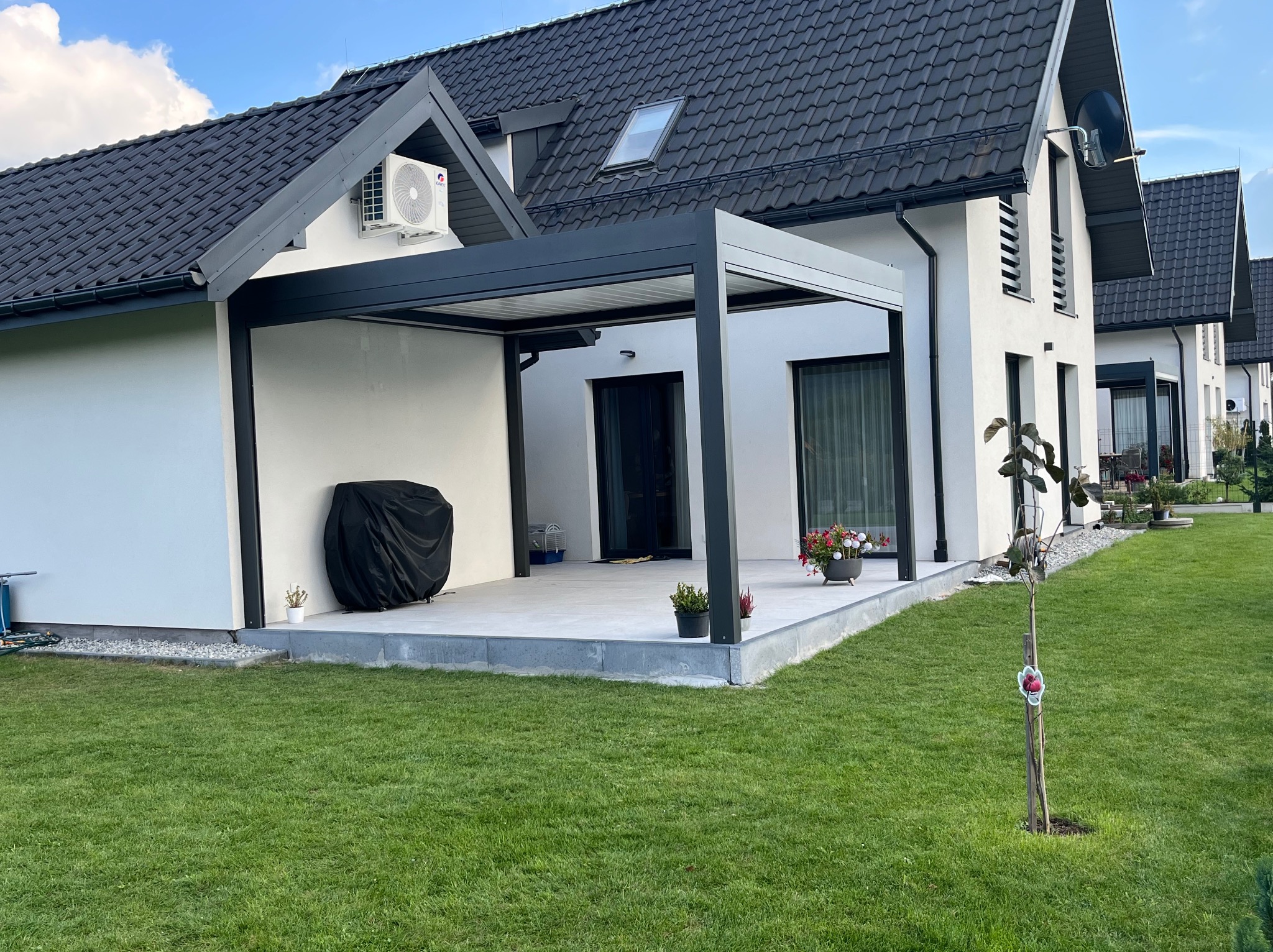 Nowoczesna, grafitowa pergola metalowa przy białym domu z czarnym dachem. Widok na zadbany trawnik i sąsiednie budynki. Minimalistyczny design.