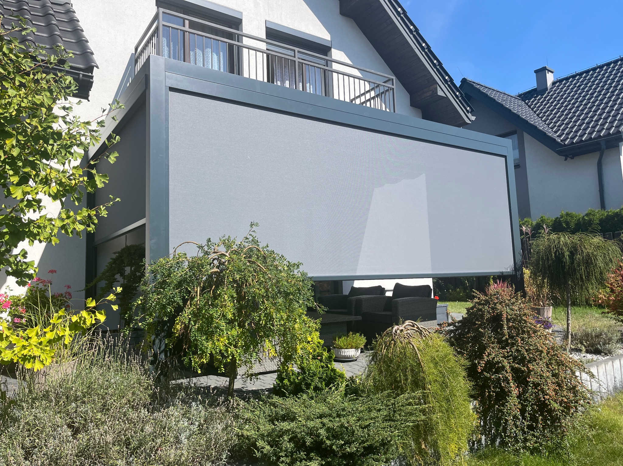 Szara, nowoczesna zabudowa balkonu z roletami przeciwsłonecznymi, widok z ogrodu. W tle biały dom z balkonem i ciemnym dachem. Uwagę przykuwają dekoracyjne krzewy.