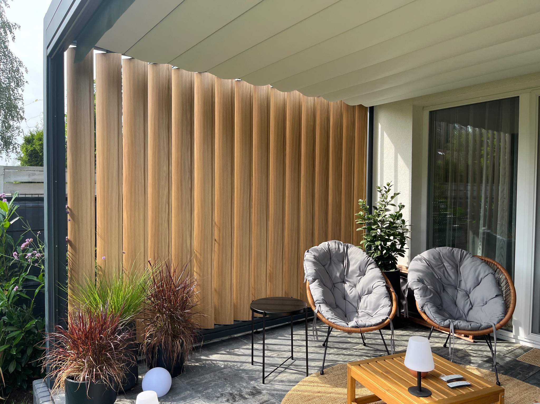 Nowoczesna pergola z drewnianymi lamelami, tworząca zacienioną przestrzeń na balkonie. Dwa fotele z poduszkami i stoliki kawowe uzupełniają aranżację.