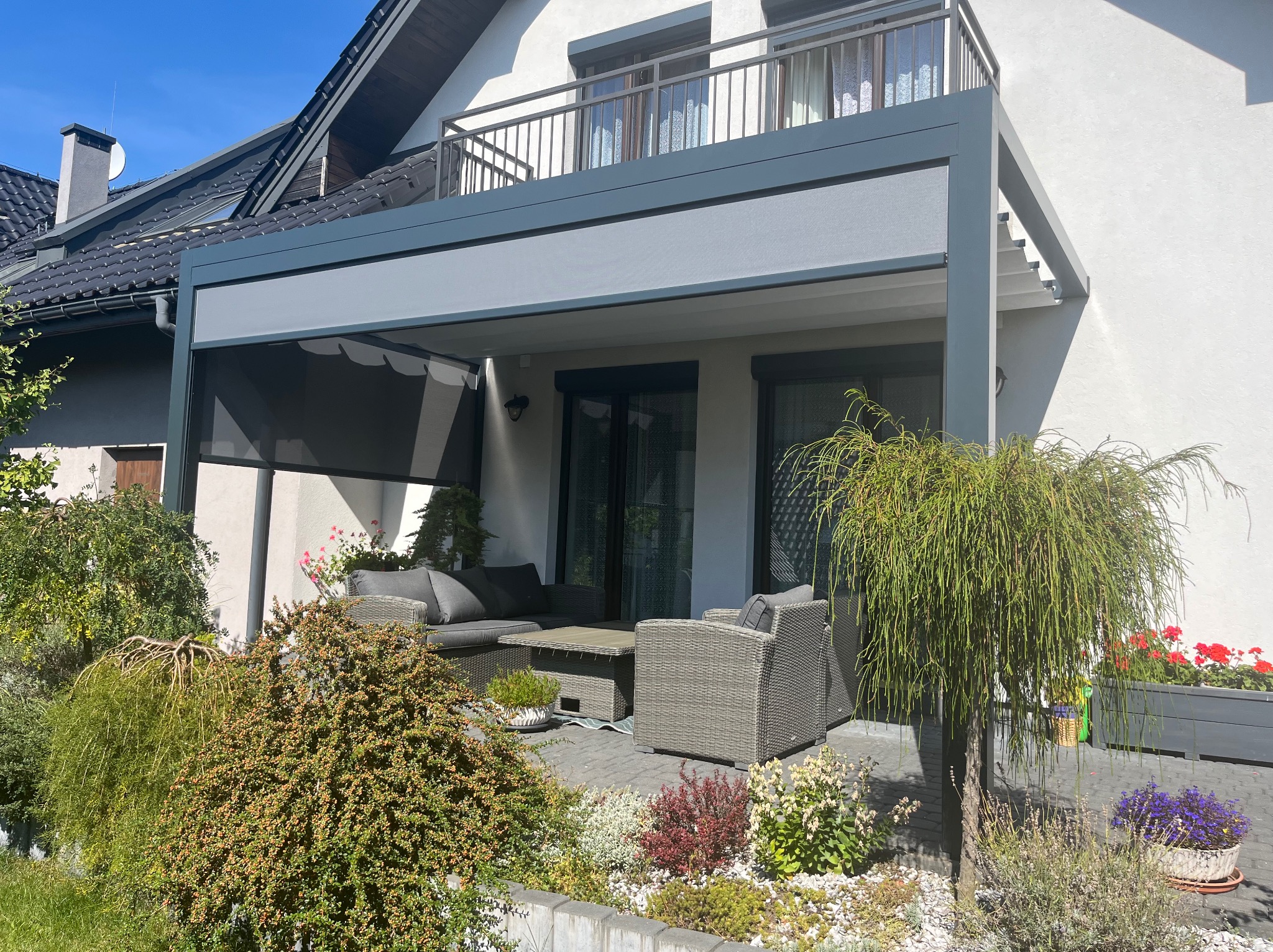 Nowoczesna, szara pergola metalowa z roletą przeciwsłoneczną, przylegająca do białego domu z balkonem. Wygodne meble ogrodowe i zadbana roślinność.