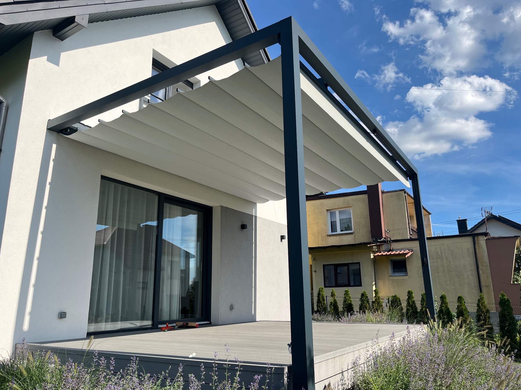 Nowoczesna pergola metalowa z rozsuwanym zadaszeniem, przylegająca do jasnego budynku z dużym oknem tarasowym. Widoczny taras z desek kompozytowych i roślinność.