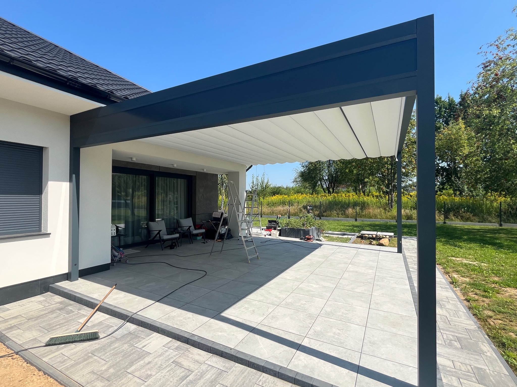 Nowoczesna, grafitowa pergola z rozsuwanym, białym zadaszeniem, przylegająca do jasnego domu. Widoczny taras z szarych płyt i fragment ogrodu w tle.