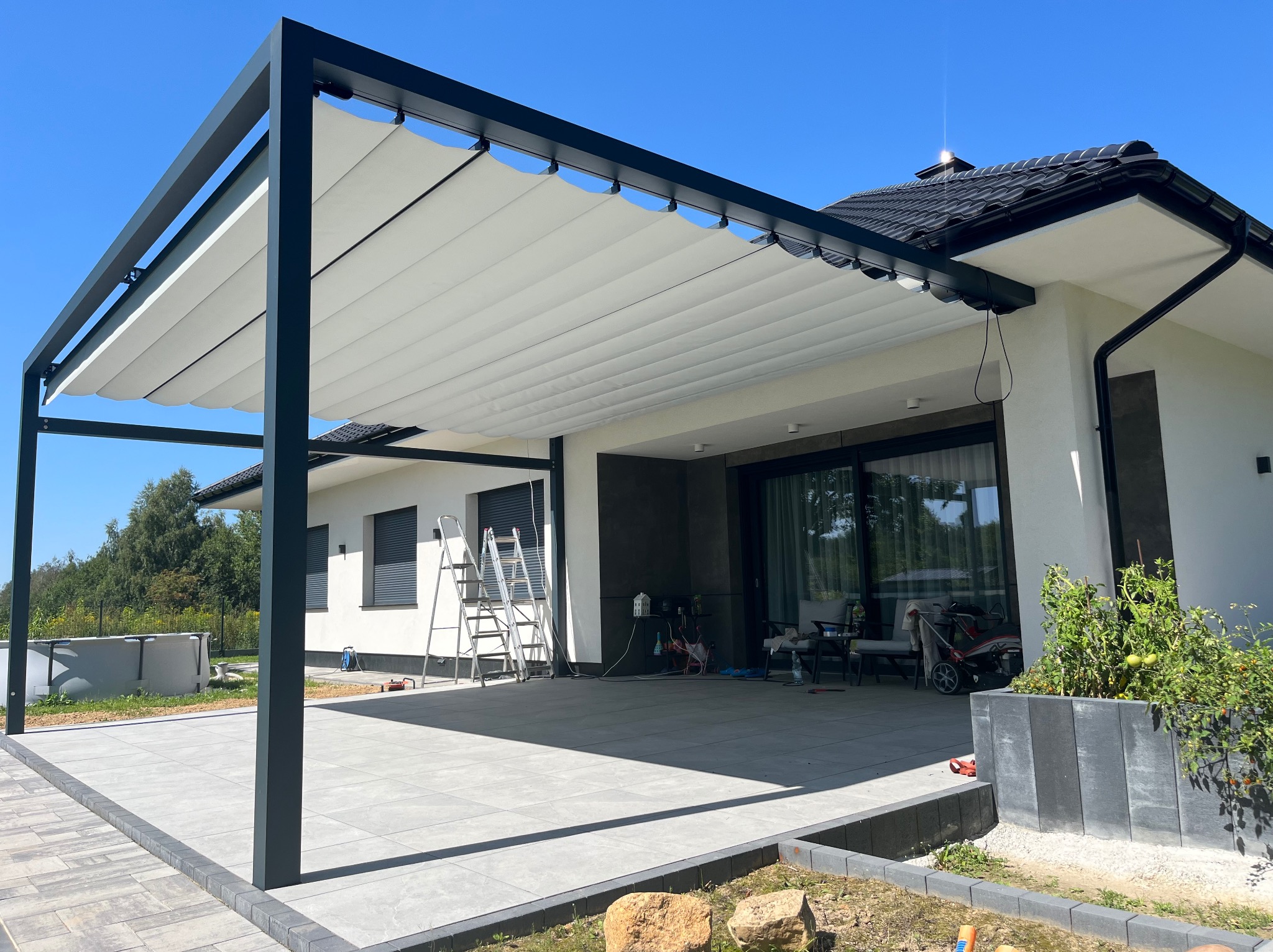 Nowoczesna, czarna pergola metalowa z białym zadaszeniem lamelowym, przylegająca do białego domu z dużym oknem tarasowym. Widoczne drabiny i narzędzia sugerują świeży montaż.