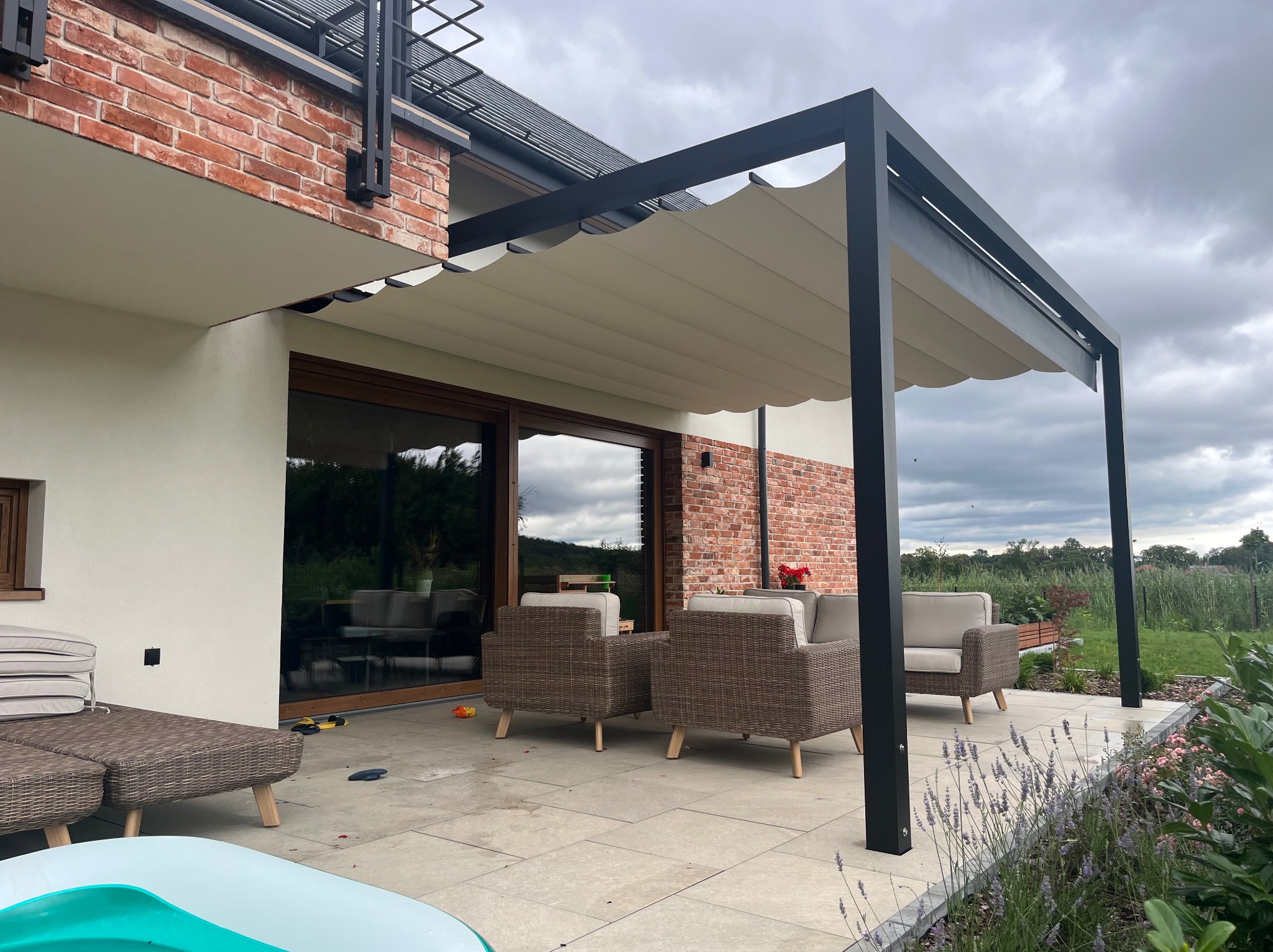 Nowoczesna, czarna pergola metalowa z beżowym zadaszeniem, przylegająca do domu z ceglaną ścianą. Na tarasie rattanowe meble ogrodowe i kwiaty. Pochmurne niebo w tle.