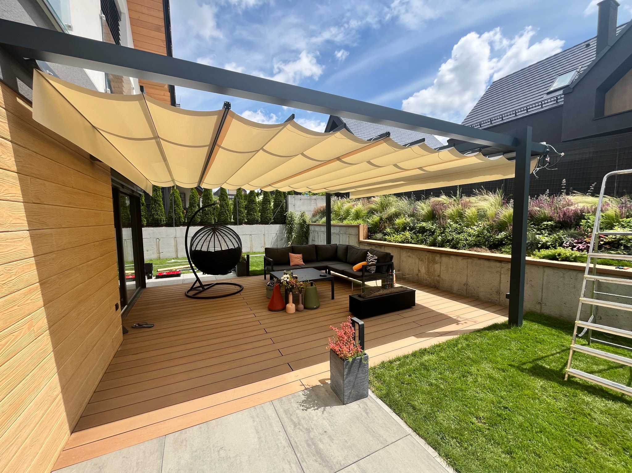 Nowoczesna pergola metalowa z beżowym, falistym zadaszeniem nad tarasem z deską kompozytową. W tle ogród i dom z czarną dachówką.