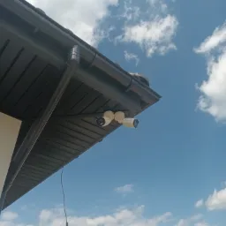 Dwie białe kamery na rogu dachu z czarną rynną, skierowane w stronę błękitnego nieba z białymi chmurami.
