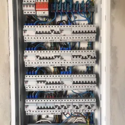 Otwarta skrzynka elektryczna z wieloma bezpiecznikami, kablami w różnych kolorach (żółty, niebieski, czarny, brązowy, zielony) i listwami zaciskowymi, zamontowana w ścianie z surowego betonu.