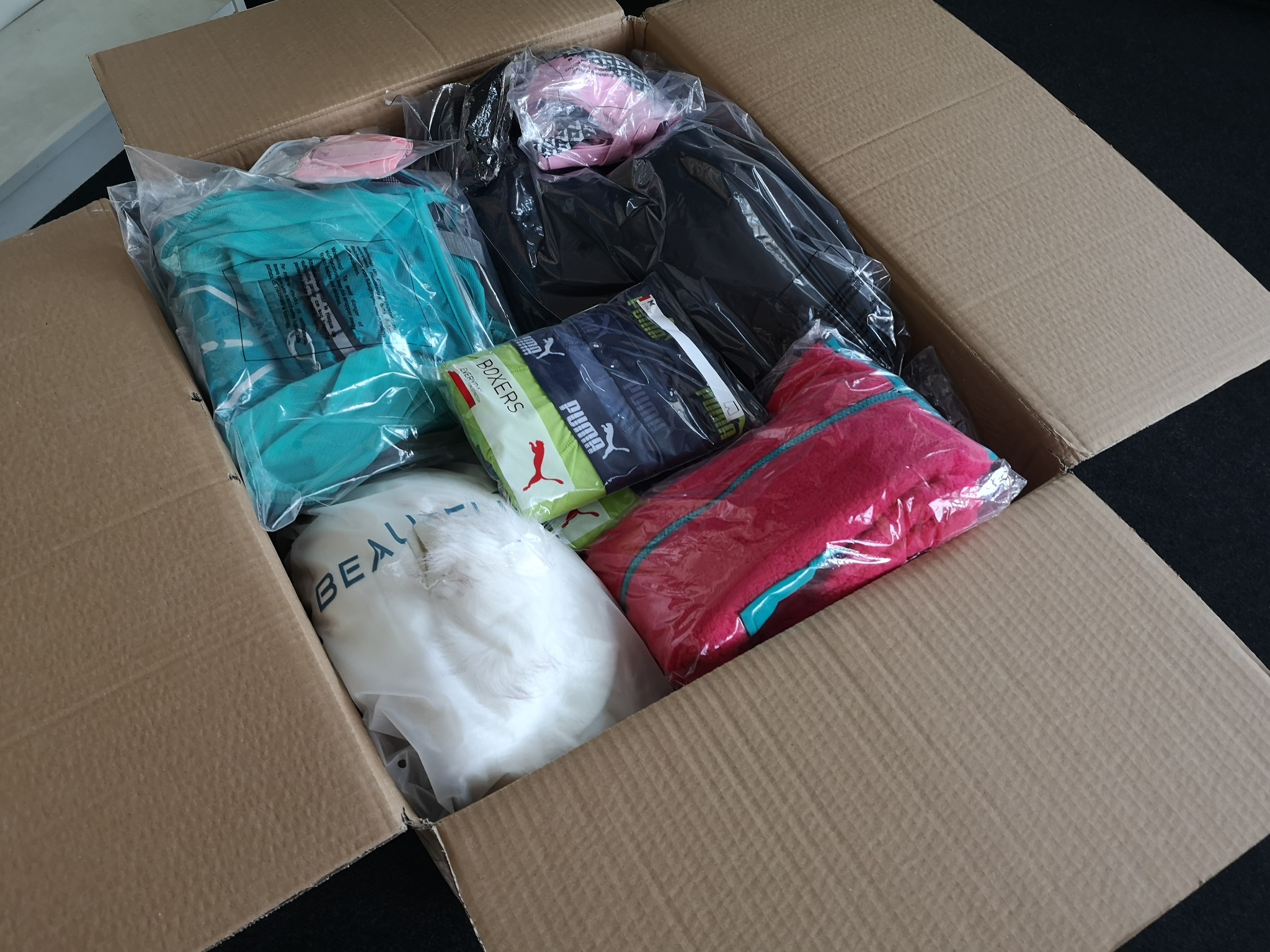 Otwarty karton wypełniony różnymi elementami odzieży w plastikowych opakowaniach, w tym bielizną marki Puma, odzieżą sportową i tekstyliami.