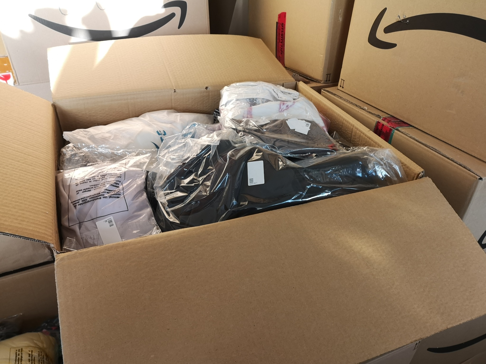 Otwarty karton z odzieżą zapakowaną w foliowe torby, widoczne etykiety i logo firmy kurierskiej na kartonach w tle.