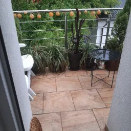 Balkon wyłożony płytkami w odcieniach beżu i brązu, z metalową balustradą ozdobioną sznurami z kulami, roślinami w donicach, białym krzesłem i czarnym stolikiem.
