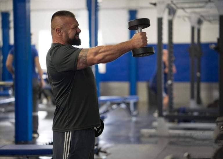Mężczyzna z tatuażem na ramieniu, w czarnej koszulce i szarych spodenkach, ćwiczy unoszenie hantli w siłowni, z grymasem wysiłku na twarzy.