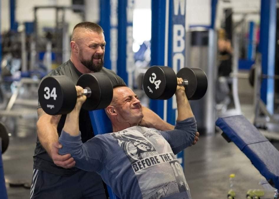 Mężczyzna wyciska sztangielki 34 kg na ławeczce pod okiem trenera w siłowni.