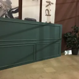 Dekoracyjna ściana w kolorze ciemnej zieleni z panelami w ramkach, ustawiona w przestrzeni publicznej z marmurową podłogą i rośliną doniczkową obok.