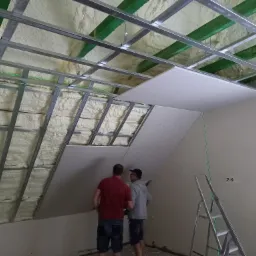 Montaż płyty gipsowo-kartonowej na poddaszu z ociepleniem, widoczne metalowe profile, pianka izolacyjna i laserowy poziom, dwóch pracowników w trakcie pracy.