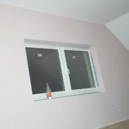 Poddasze po malowaniu na różowo, białe okno z butelką w środku, widoczne skosy i fragment podłogi.