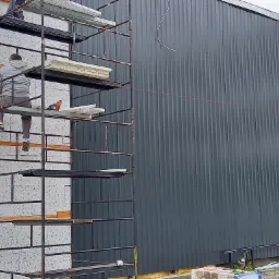 Pracownik na rusztowaniu montuje izolację termiczną na ścianie budynku z blachy trapezowej, widoczna izolacja styropianowa i narzędzia.