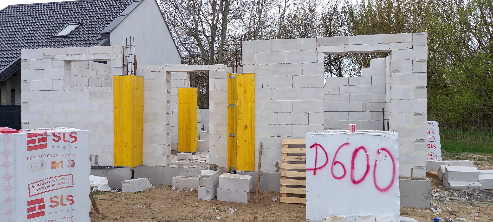 Budowa domu jednorodzinnego: ściany z białych bloczków, żółte szalunki okienne, zbrojenia pionowe, paleta i opakowania bloczków SLS z napisem 'P600'