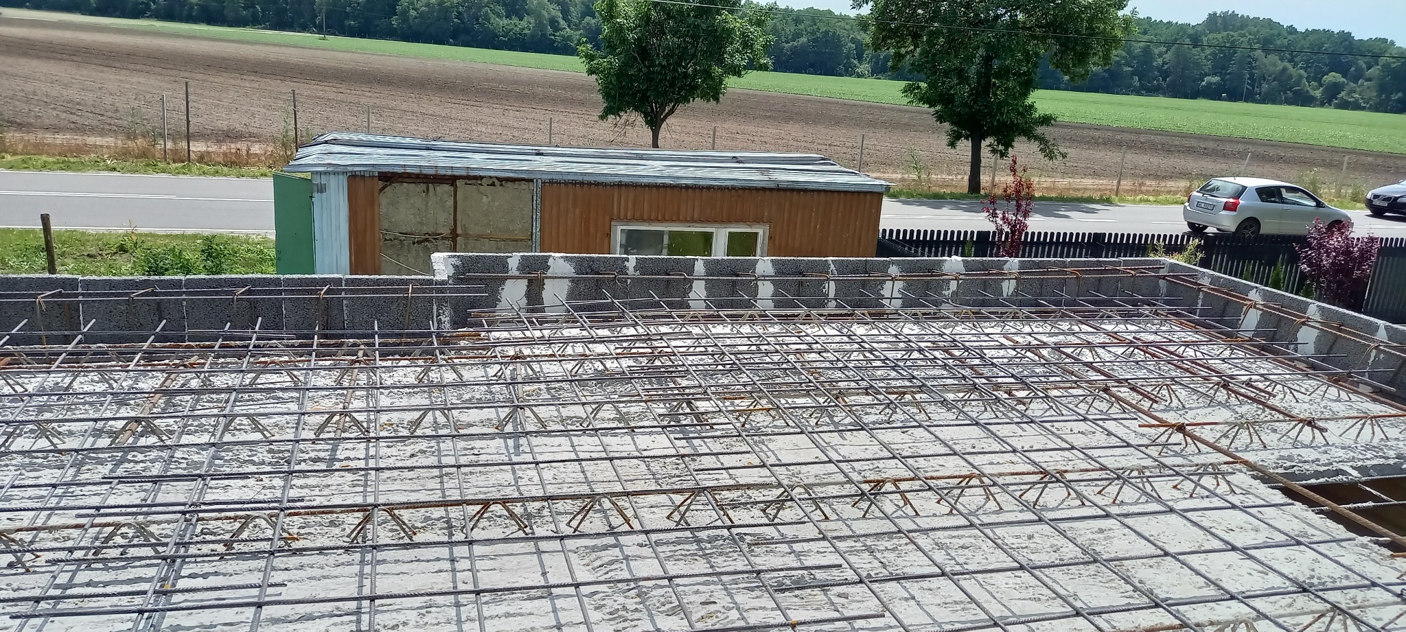 Zbrojenie fundamentów z betonu na budowie domu jednorodzinnego, widok z góry na siatkę z prętów stalowych, w tle budynek gospodarczy i droga.
