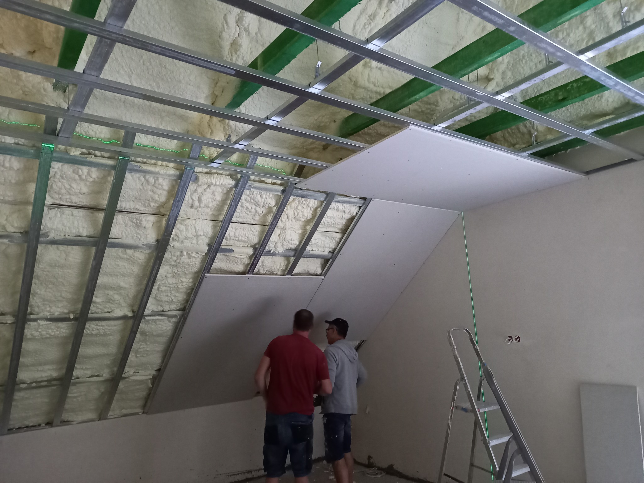 Montaż płyty gipsowo-kartonowej na poddaszu z ociepleniem, widoczne metalowe profile, pianka izolacyjna i laserowy poziom, dwóch pracowników w trakcie pracy.