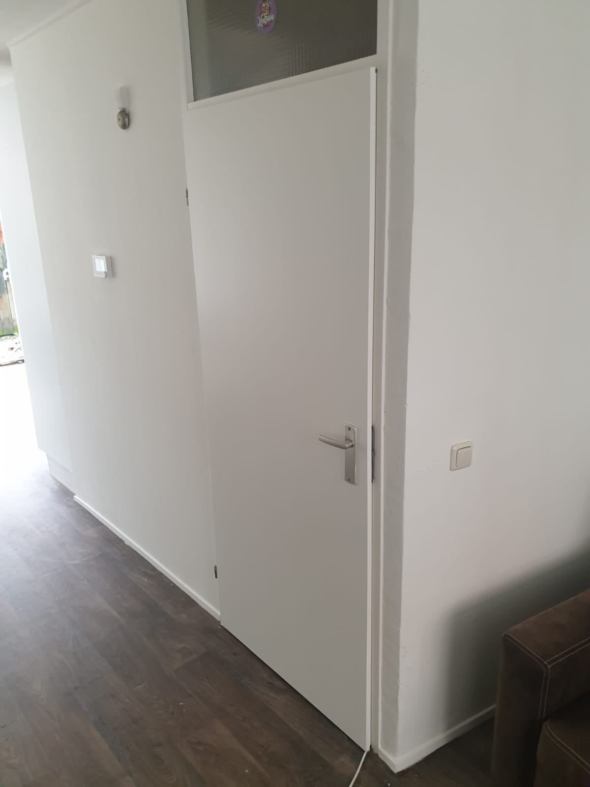 Białe drzwi wewnętrzne z szybą u góry, zamontowane w białej ścianie, widoczny fragment brązowej podłogi i narożnik sofy.