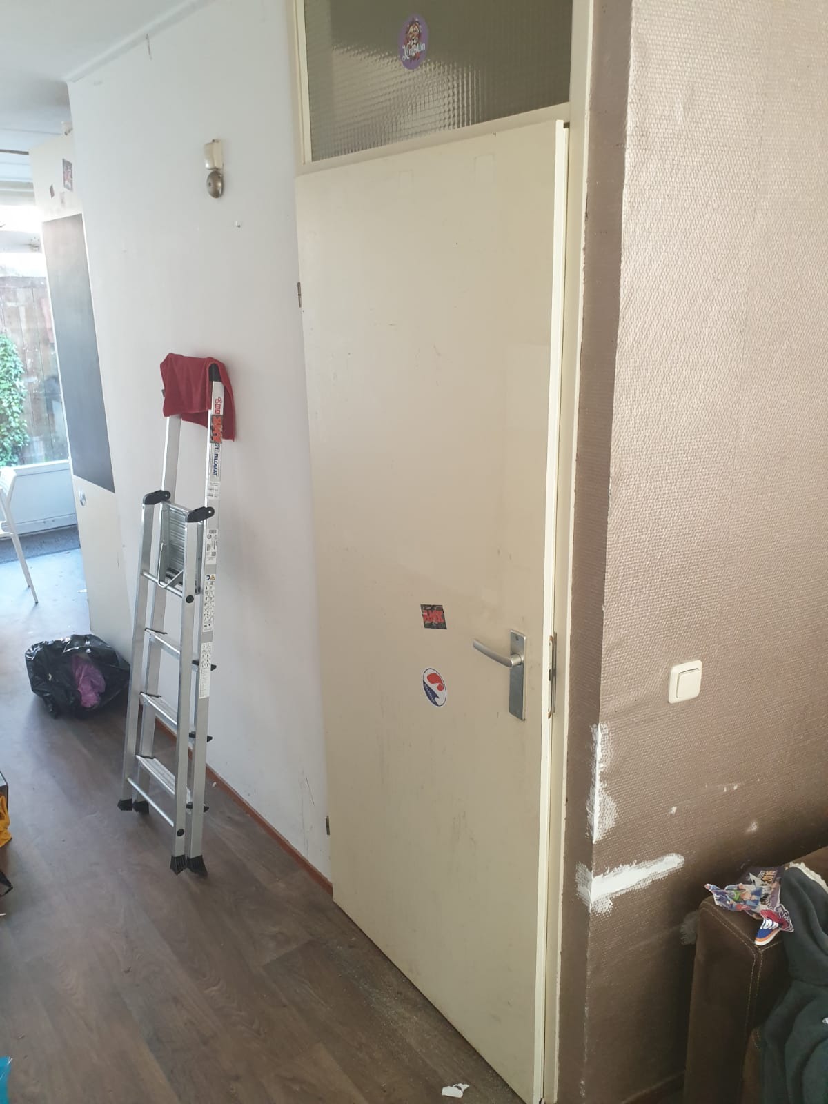 Wnętrze mieszkania w trakcie odświeżania: drabina oparta o ścianę, biała, stara, lekko zabrudzona futryna drzwi z naklejkami, fragment ściany po szpachlowaniu, widoczne przygotowania do malowania.