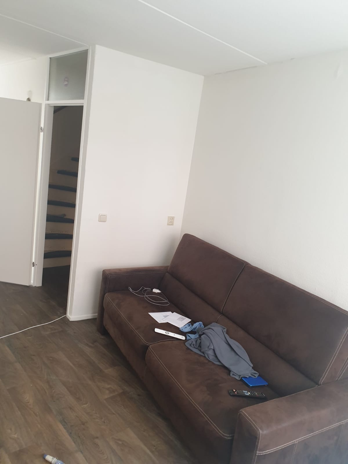 Widok wnętrza pokoju z brązową kanapą, białymi ścianami po odświeżeniu i podłogą z paneli imitujących drewno, z otwartymi drzwiami prowadzącymi do schodów.