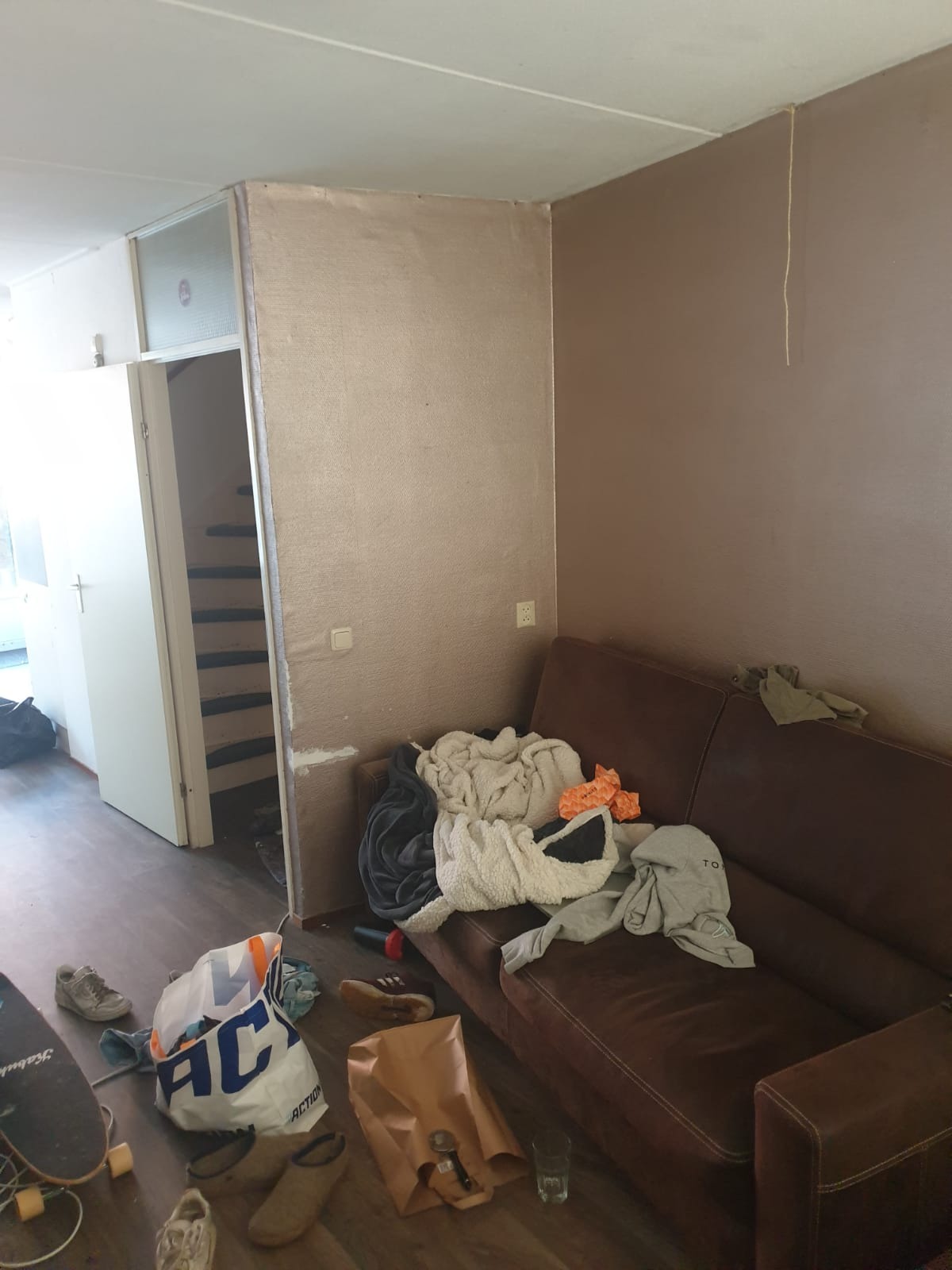 Pokój w trakcie remontu: świeżo pomalowana ściana w odcieniach beżu, brązowa kanapa z porozrzucanymi ubraniami, podłoga z deskami i torba na zakupy leżąca obok deski do skateboardingu.