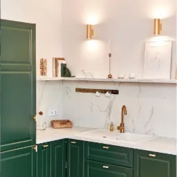 Kącik kuchenny z zielonymi szafkami, marmurowym blatem i złotą armaturą. Nad zlewem półka z dekoracjami i uchwytami na kubki.