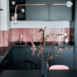 LBB Nieruchomości Sp. z o.o. - Nowoczesna kuchnia z ciemnymi szafkami, różową mozaiką na ścianie i designerską lampą wiszącą nad stołem z wazonem z kwitnącymi gałązkami.