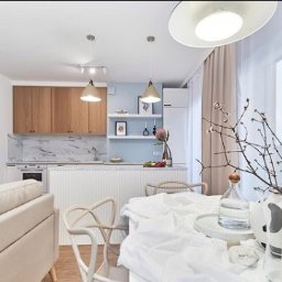 LBB Nieruchomości Sp. z o.o. - Jasne wnętrze z otwartą kuchnią i jadalnią, drewniane szafki kuchenne z marmurowym blatem, biały stół nakryty białym obrusem, minimalistyczne dekoracje, okrągłe lustra na ścianie.