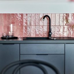 LBB Nieruchomości Sp. z o.o. - Wrocław: Nowoczesna kuchnia z szarymi szafkami i różowymi, błyszczącymi płytkami wertykalnymi nad blatem, uzupełniona czarną armaturą i szklanym dzbankiem.