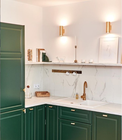 Kącik kuchenny z zielonymi szafkami, marmurowym blatem i złotą armaturą. Nad zlewem półka z dekoracjami i uchwytami na kubki.