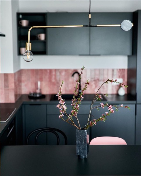 Nowoczesna kuchnia z ciemnymi szafkami, różową mozaiką na ścianie i designerską lampą wiszącą nad stołem z wazonem z kwitnącymi gałązkami.