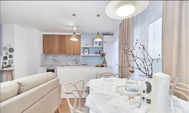 Jasne wnętrze z otwartą kuchnią i jadalnią, drewniane szafki kuchenne z marmurowym blatem, biały stół nakryty białym obrusem, minimalistyczne dekoracje, okrągłe lustra na ścianie.