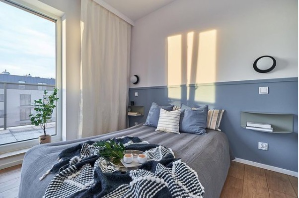 Jasna sypialnia w błękitach z drewnianą podłogą, łóżko z szarą narzutą i dekoracyjną pledem, stolik z filiżankami kawy oraz widokiem na balkon z rośliną w doniczce.