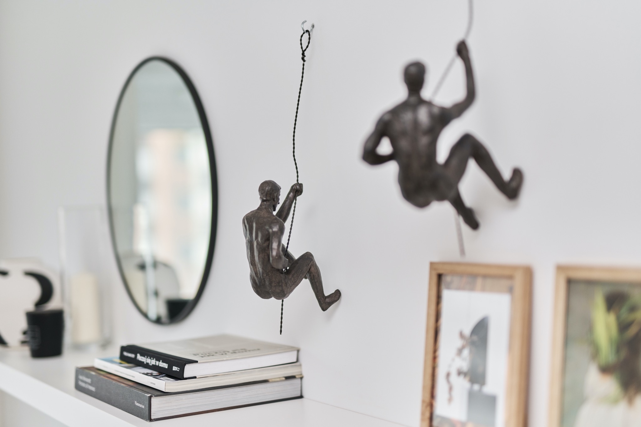 Dekoracja ścienna z figurkami wspinaczy zawieszonymi na białej ścianie nad półką z książkami i owalnym lustrem.