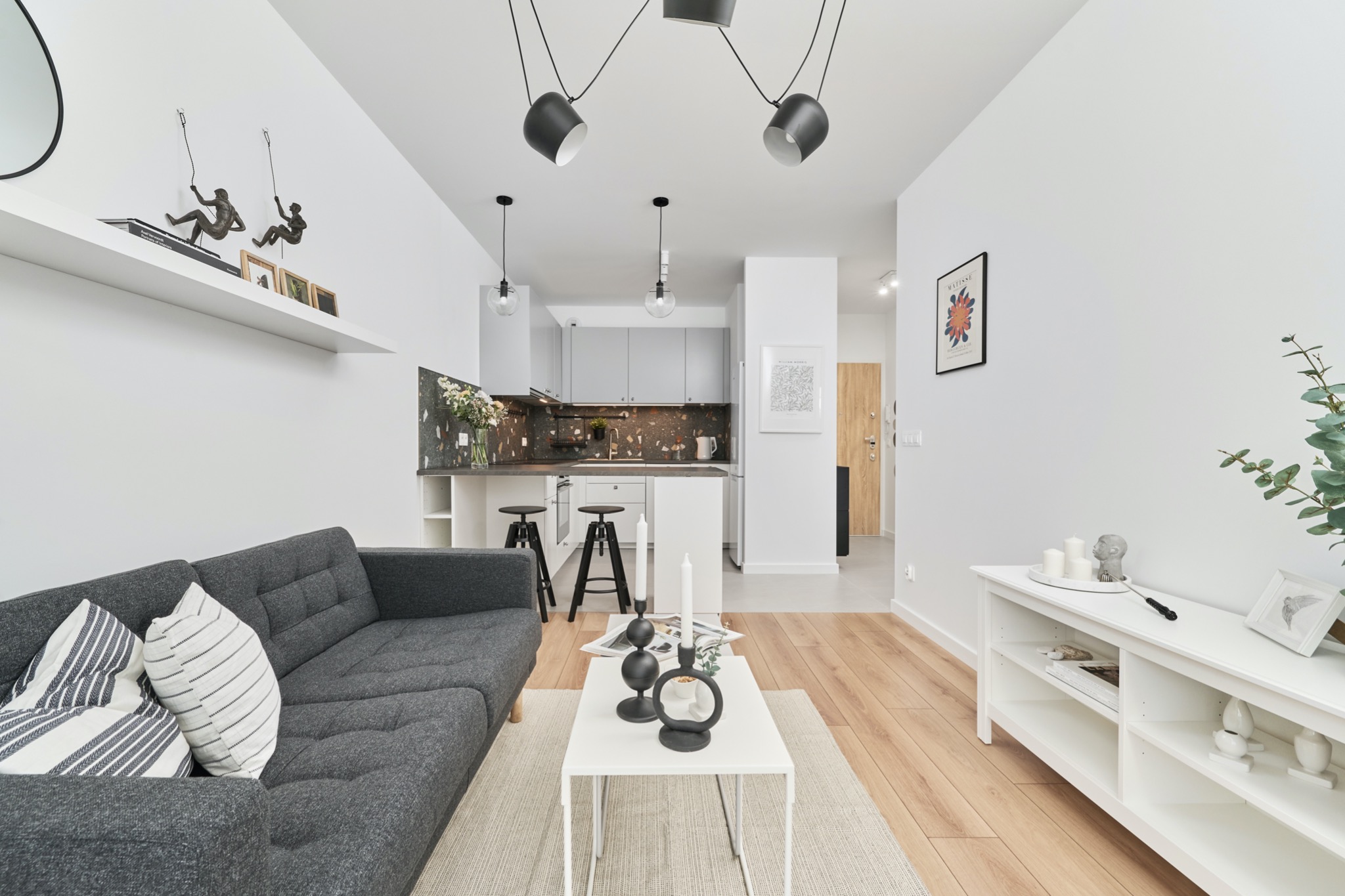 Jasne wnętrze mieszkania z szarym narożnikiem, białym stolikiem kawowym z dekoracjami i otwartą kuchnią w tle, oświetlone trzema lampami sufitowymi.