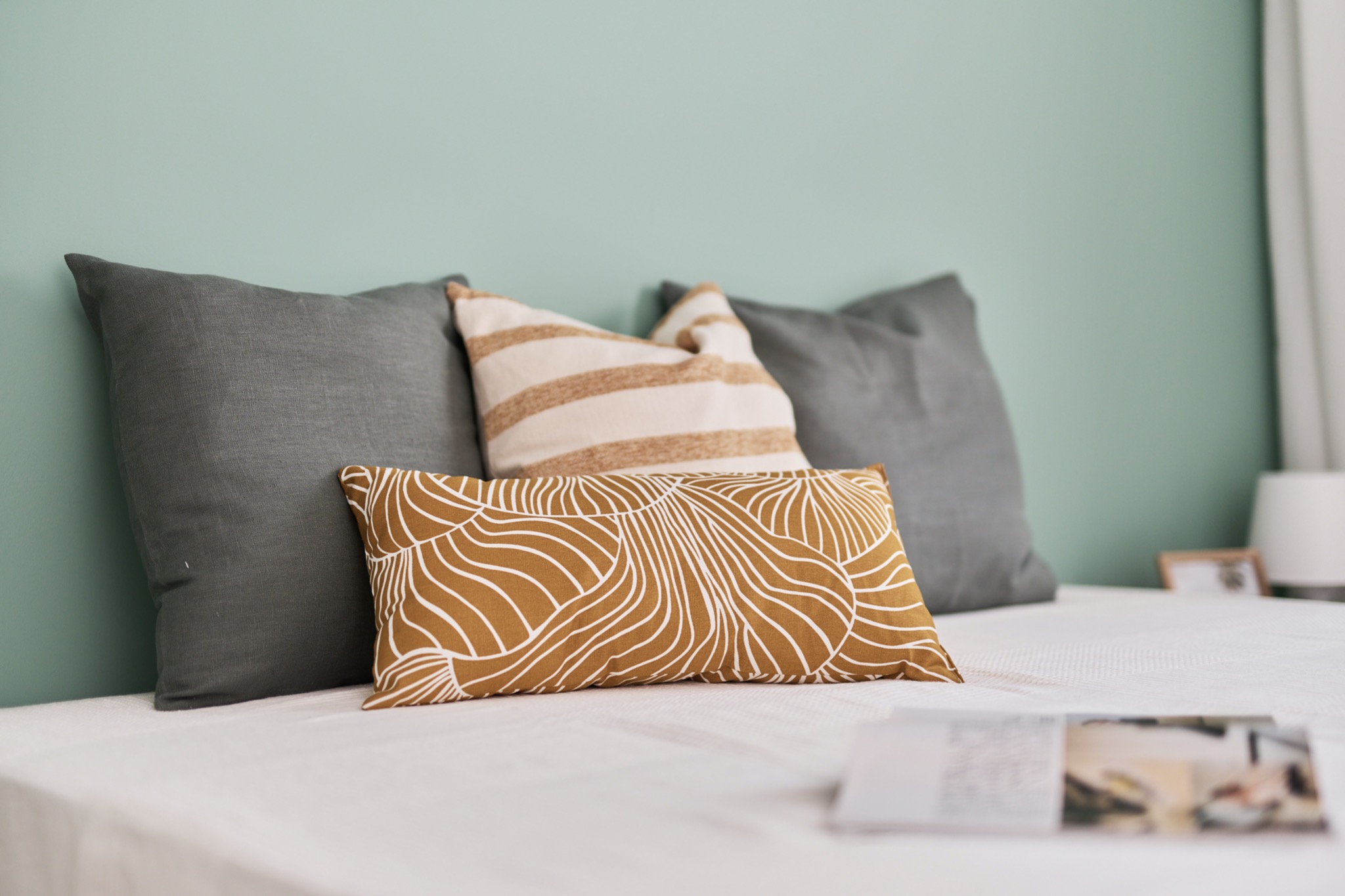 Łóżko z szarymi poduszkami, poduszką w beżowe paski i poduszką w musztardowe wzory na tle miętowej ściany.