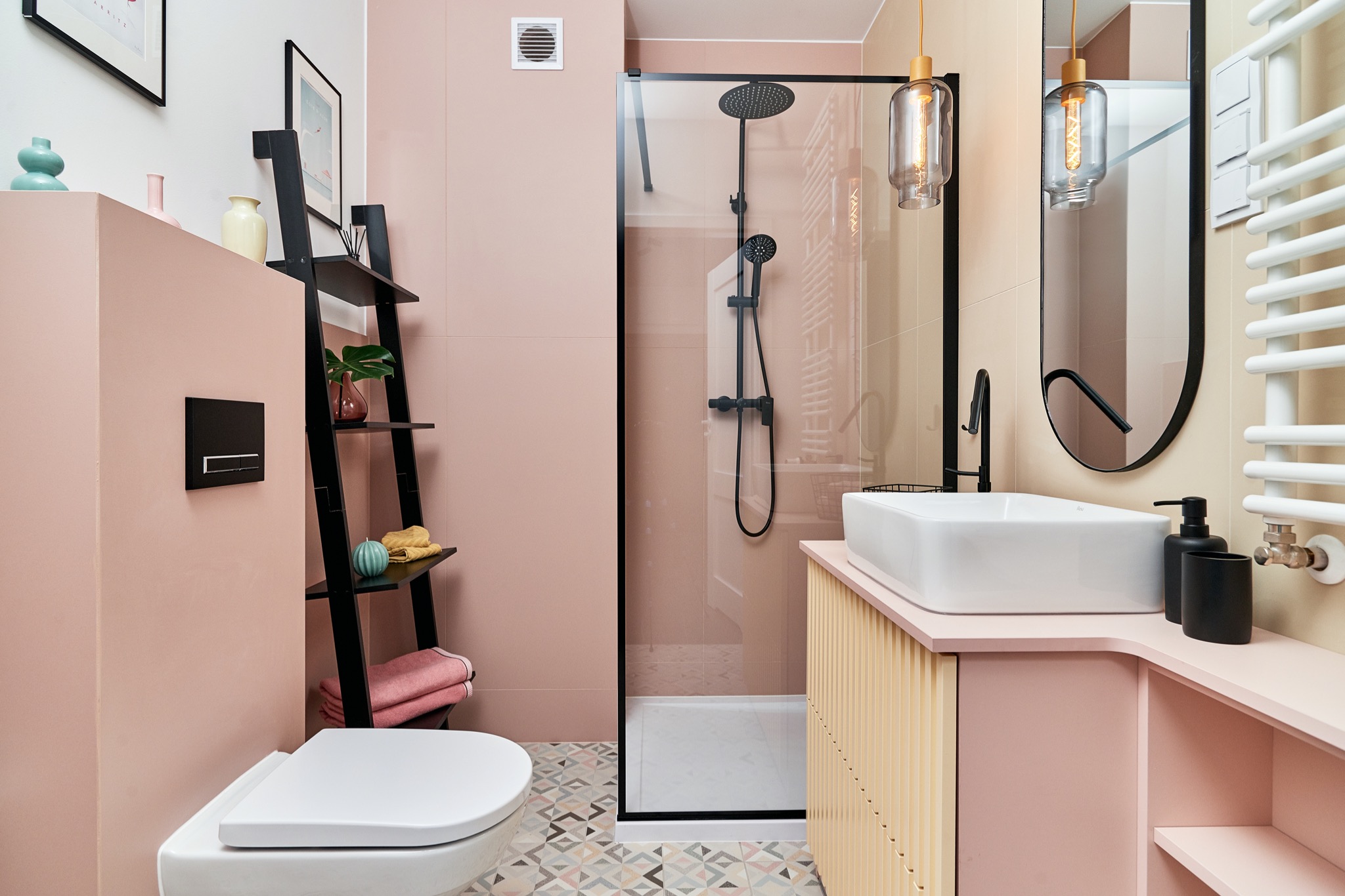 Różowa łazienka z czarnymi akcentami: kabina prysznicowa typu walk-in, umywalka nablatowa, drabina-regał z dekoracjami, wzorzysta podłoga.