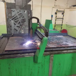 Automatyczna wycinarka plazmowa w trakcie pracy, iskry widoczne podczas cięcia metalowego arkusza na stole roboczym, w tle ściany warsztatu i metalowe elementy.