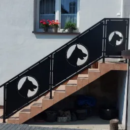 Zewnętrzne schody z metalową balustradą ozdobioną motywem psiej głowy w okręgu, prowadzące do piwnicy budynku z oknem udekorowanym kwiatami.